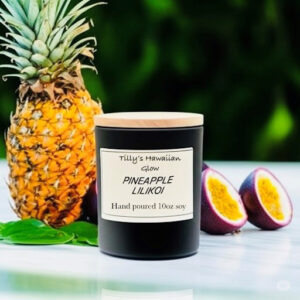 Tilly's Hawaiian Glow Pineapple Lilikoi 10 oz hand-poured black soy candle with wooden lid, next to a whole pineapple and sliced passion fruits.