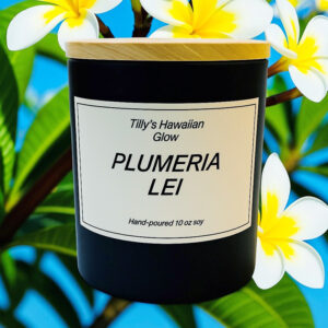 Tilly's Hawaiian Glow Plumeria Lei 10 oz hand-poured black soy candle with wooden lid, surrounded by white and yellow plumeria flowers against a blue sky.
