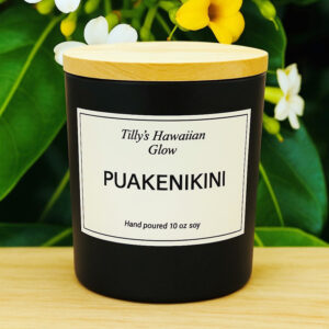 Tilly's Hawaiian Glow Puakenikini 10 oz hand-poured black soy candle with wooden lid, surrounded by white and yellow Puakenikini flowers.